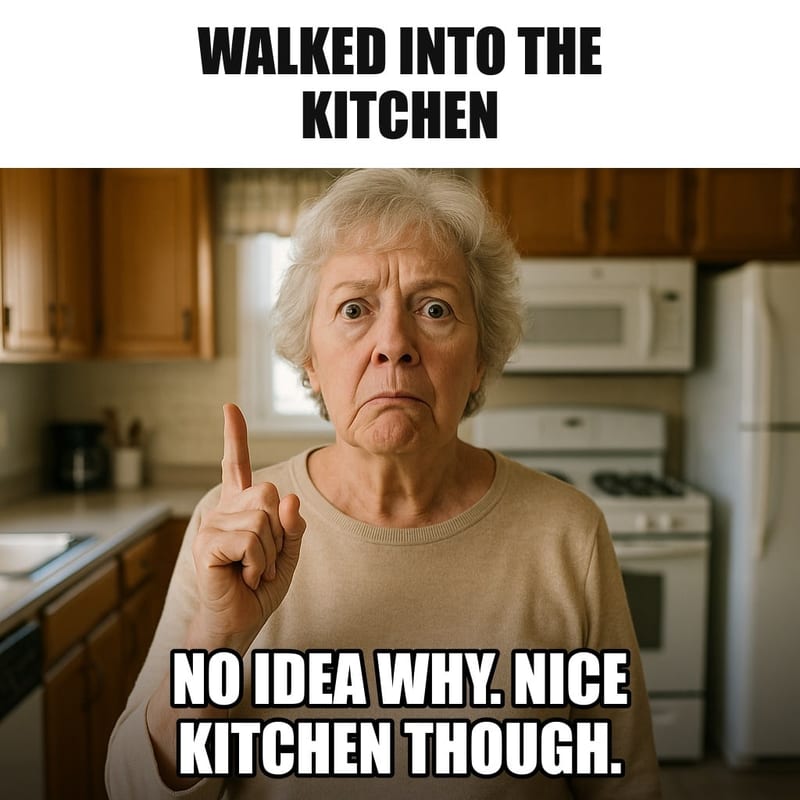 A senior woman standing in the middle of her kitchen with a puzzled, blank look, one finger raised as if about to remember something. Caption: "Walked into the kitchen No idea why. Nice kitchen though."